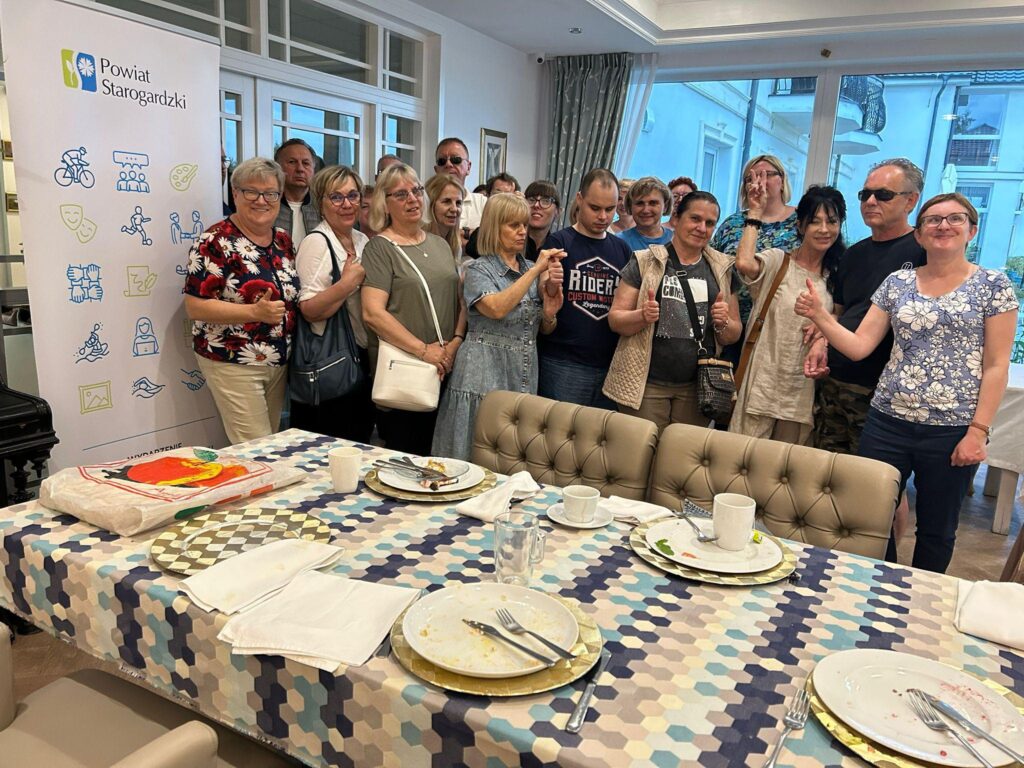 Grupa około piętnastu osób stoi razem w pomieszczeniu, uśmiechając się do aparatu. W tle po lewej stronie jest pionowy baner z napisem "Powiat Starogardzki" i kilkoma kolorowymi ikonami przedstawiającymi różne aktywności, takie jak jazda na rowerze, teatr, malarstwo, sport i inne. Osoby są w różnym wieku, większość to kobiety, a dwie osoby noszą okulary przeciwsłoneczne. Przed nimi znajduje się stół nakryty geometrycznym obrusie w odcieniach niebieskiego, szarego i beżu. Na stole są puste talerze, filiżanki i sztućce, co sugeruje, że grupa właśnie skończyła posiłek lub spotkanie przy kawie. W tle po prawej stronie widać duże okna, przez które widać budynek i balkon. Atmosfera wydaje się przyjazna i radosna.