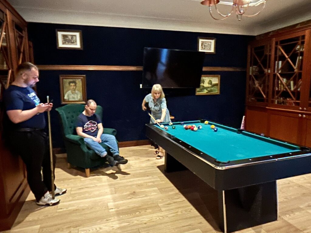 Pokój z bilardem, w którym znajdują się trzy osoby. Po lewej stronie stoi mężczyzna w ciemnej koszulce i jasnych butach, trzymający kij bilardowy i patrzący na telefon. Obok niego na zielonym fotelu siedzi drugi mężczyzna w ciemnej koszulce i jasnych spodniach, z rękami na kolanach, patrzący w dół. Po prawej stronie przy stole bilardowym pochylona jest kobieta o blond włosach, ubrana w jasną sukienkę, trzymająca kij bilardowy i przygotowująca się do uderzenia. W tle na ścianie wiszą obrazy i duży telewizor. Meble są drewniane, a podłoga jasna, drewniana. Ściany są ciemne, prawdopodobnie granatowe.