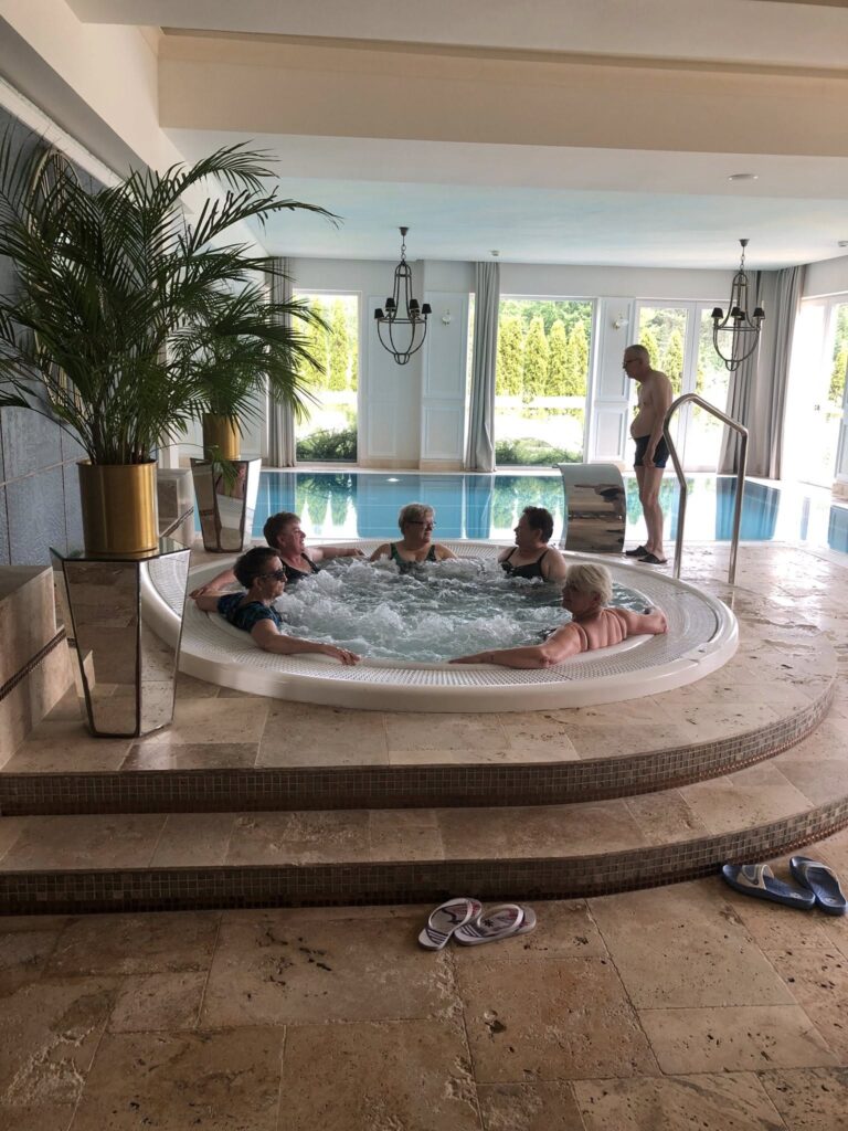 Zdjęcie przedstawia pięć osób siedzących w okrągłym jacuzzi w pomieszczeniu z dużym basenem w tle. Jacuzzi jest wbudowane w podwyższenie z kamiennymi schodami prowadzącymi do niego. W jacuzzi znajdują się cztery kobiety i jeden mężczyzna, wszyscy w strojach kąpielowych. W tle, przy basenie, stoi starszy mężczyzna w kąpielówkach i klapkach. Po obu stronach jacuzzi stoją duże donice z wysokimi zielonymi roślinami. Pomieszczenie jest jasne, z dużymi oknami, przez które widać zieleń na zewnątrz. Na podłodze przed schodami leżą dwie pary klapek. Wnętrze wygląda na nowoczesne i zadbane.