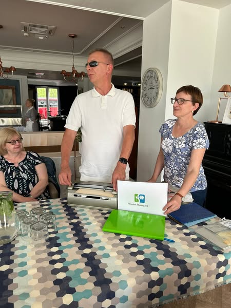 Zdjęcie przedstawia trzy osoby stojące i siedzące przy stole w jasnym pomieszczeniu. Mężczyzna w środku ma krótkie włosy, nosi okulary przeciwsłoneczne i białą koszulkę polo. Po jego prawej stronie stoi kobieta w krótkich włosach, ubrana w niebieską bluzkę w białe kwiaty i okulary. Po lewej stronie siedzi inna kobieta z blond włosami, ubrana w ciemną bluzkę z jasnym wzorem. Na stole leży zielony folder z napisem "Powiat Starogardzki" oraz kilka szklanek i dzbanek z wodą. W tle widać zegar na ścianie i fragment fortepianu. Pomieszczenie jest jasne, z dużym oknem, przez które widać czerwony pojazd na zewnątrz.