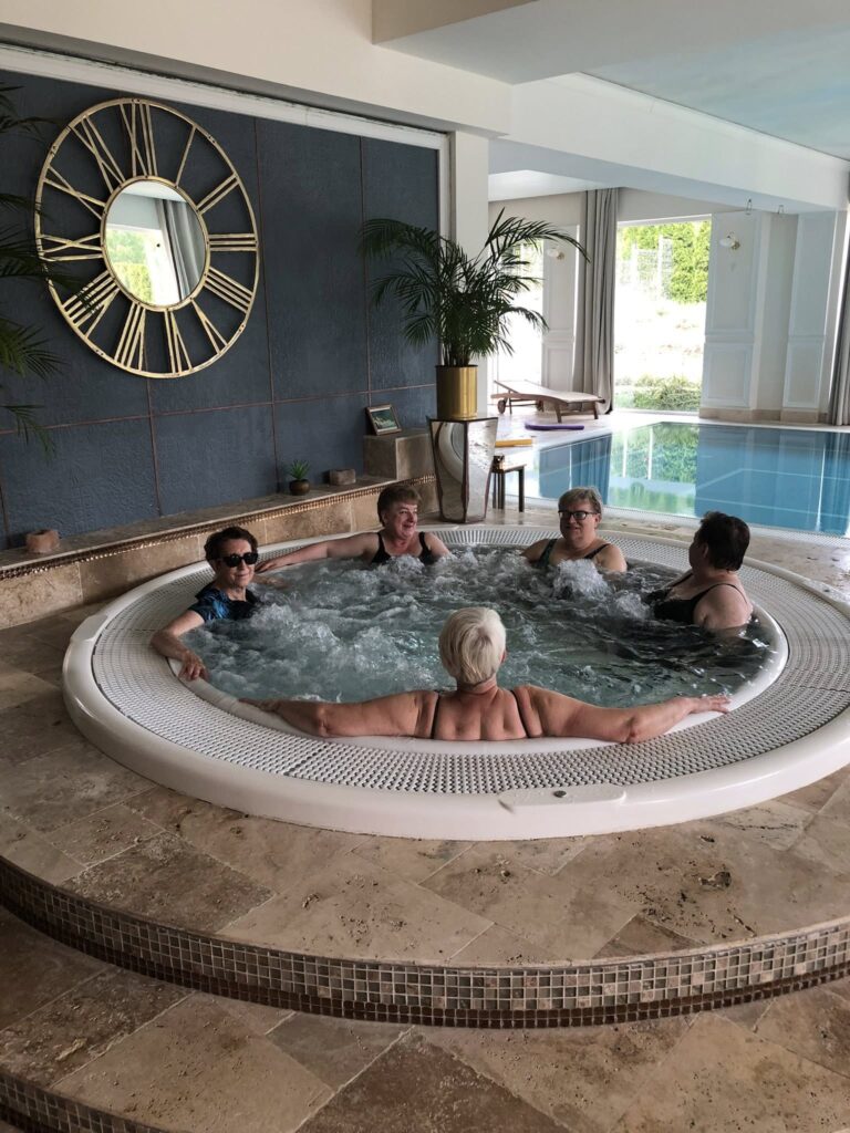 Zdjęcie przedstawia pięć kobiet siedzących w okrągłym jacuzzi w pomieszczeniu z basenem. Kobiety są w kostiumach kąpielowych, jedna z nich ma krótkie jasne włosy i jest zwrócona tyłem do aparatu, pozostałe cztery są zwrócone do przodu i uśmiechają się. W tle widać basen, duże okna wpuszczające światło dzienne oraz ścianę z dużym, ozdobnym zegarem z rzymskimi cyframi i lustrem w środku. W pomieszczeniu są też rośliny doniczkowe i meble. Podłoga jest wyłożona jasnym kamieniem lub płytkami. Cała scena wygląda na relaksującą i przyjemną.