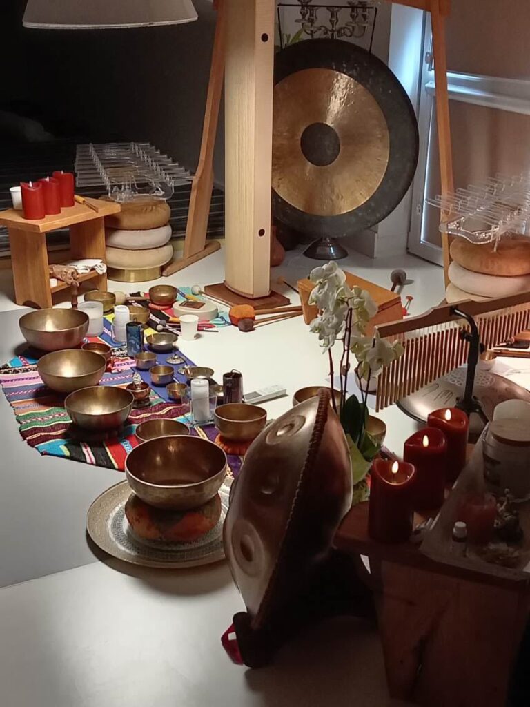 Zdjęcie przedstawia pomieszczenie z różnorodnymi instrumentami muzycznymi i przedmiotami do medytacji lub relaksacji. Na pierwszym planie widoczna jest duża misa dźwiękowa umieszczona na poduszce, obok niej kilka mniejszych mis. Na stole obok stoi kilka czerwonych świec, które są zapalone. W tle znajduje się duży gong zawieszony na drewnianej ramie oraz różne inne instrumenty perkusyjne, takie jak dzwonki, małe misy i pałeczki. Na podłodze leży kolorowy, wzorzysty dywanik, na którym poustawiane są kolejne misy i przedmioty. W pomieszczeniu panuje ciepła, przytulna atmosfera, sprzyjająca wyciszeniu i medytacji.