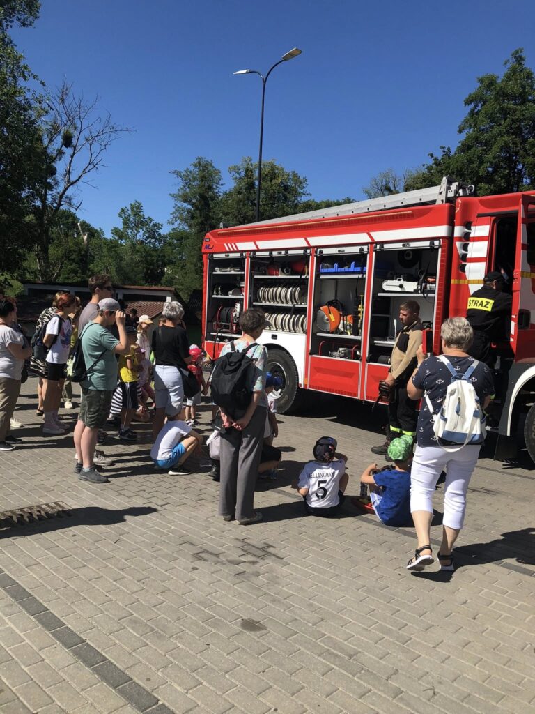 Wóz strażacki otoczony przez grupę ludzi, głównie dzieci i dorosłych. Wóz ma otwarte boczne schowki, w których widać różne sprzęty strażackie, takie jak węże, narzędzia i gaśnice. Kilkoro dzieci siedzi na ziemi przed pojazdem, a reszta osób stoi wokół, patrząc na strażaków i sprzęt. Dwóch strażaków w mundurach z napisem "STRAŻ" prezentuje wyposażenie wozu. Pogoda jest słoneczna, niebo bezchmurne, a cała scena rozgrywa się na wybrukowanym placu, w otoczeniu zielonych drzew. Wygląda to na pokaz lub spotkanie edukacyjne ze strażakami.