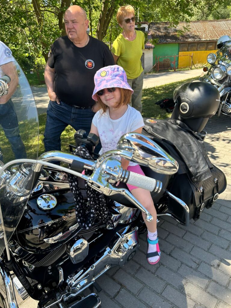 Dziewczynka siedząca na dużym, czarnym motocyklu. Ma na sobie różowy kapelusz z uśmiechniętą buźką, okulary przeciwsłoneczne, białą koszulkę i różowe spodenki. Na nogach ma kolorowe sandały i skarpetki. Obok niej stoi starszy mężczyzna w czarnej koszulce z naszywką oraz starsza kobieta w jasnozielonej bluzce i okularach przeciwsłonecznych. W tle widać jeszcze jeden motocykl, drzewa oraz kolorowy budynek z blaszanym dachem. Cała scena odbywa się w słoneczny dzień na chodniku.
