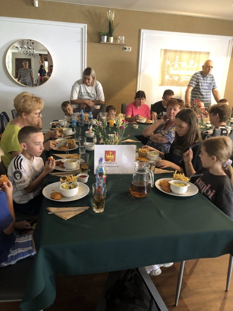 Grupa dzieci i dorosłych siedzących przy dużym stole w restauracji. Na stole leży ciemnozielony obrus, a na środku stoi dzbanek z napojem, kilka butelek wody, szklanki oraz wazonik z różowymi kwiatami. Każdy ma przed sobą talerz z jedzeniem – widać panierowane kotlety, frytki i surówki. Na stole stoi też tabliczka z napisem „GMINA MIEJSKA PRUSZCZ GDAŃSKI” oraz herbem miasta. W tle widać kolejne osoby siedzące przy innym stole, a na ścianie wisi okrągłe lustro, w którym odbija się kilka osób. Atmosfera jest rodzinna i swobodna, wygląda na wspólny posiłek, być może podczas wycieczki lub kolonii.