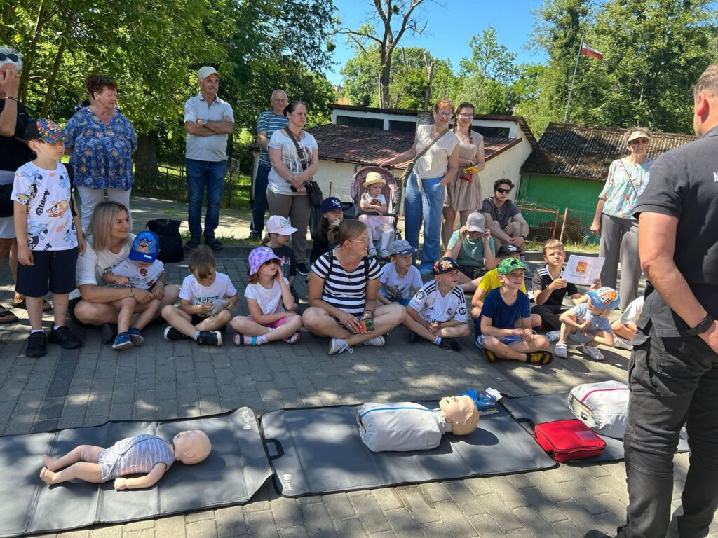 Grupa dzieci i dorosłych siedzących i stojących na zewnątrz, na chodniku w cieniu drzew. Przed nimi na matach leżą trzy fantomy do nauki pierwszej pomocy: jeden niemowlęcy i dwa większe, przypominające dorosłych. Obok fantomów znajdują się torby ratownicze. W tle widać budynki gospodarcze z zielonymi i brązowymi ścianami oraz polską flagę na maszcie. Dzieci i dorośli uważnie patrzą na osobę prowadzącą pokaz, która stoi po prawej stronie zdjęcia tyłem do aparatu. Pogoda jest słoneczna, a większość osób ma letnie ubrania i czapki z daszkiem.