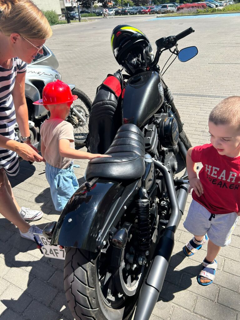 Czarny motocykl zaparkowany na chodniku, wokół którego stoją trzy osoby: kobieta w okularach przeciwsłonecznych i w sukience w paski, mały chłopiec w czerwonym kasku strażackim oraz drugi chłopiec w czerwonej koszulce i jasnych spodenkach. Kobieta pochyla się w stronę chłopca z kaskiem, który dotyka motocykla. Drugi chłopiec patrzy w stronę motocykla i ma na sobie sandały. Na motocyklu leży kask motocyklowy z żółtymi elementami oraz czerwono-czarna kurtka. W tle widać drugi motocykl, zaparkowane samochody i fragment zieleni. Jest słoneczny dzień.