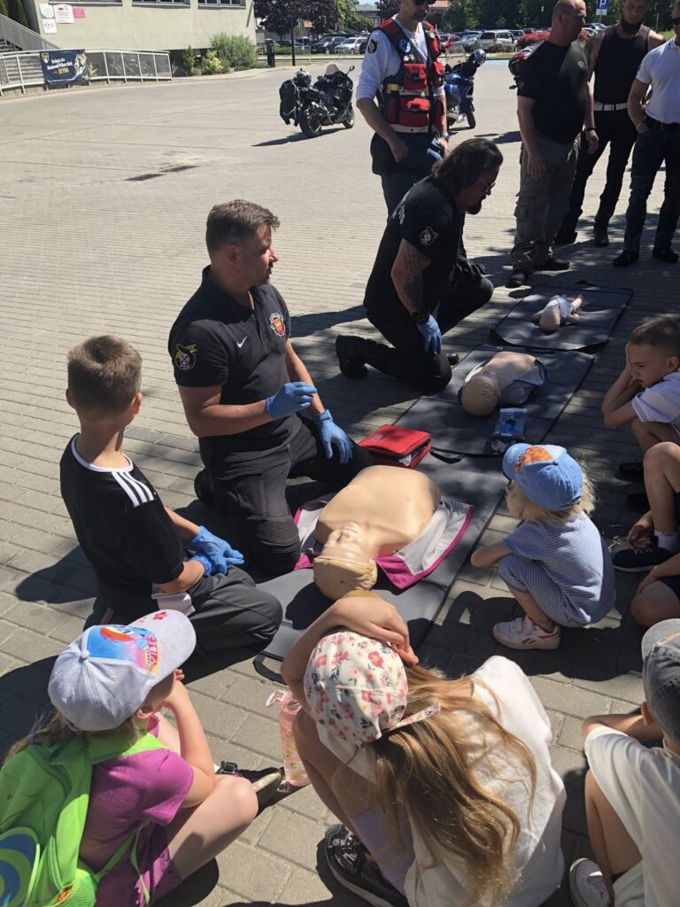 Pokaz pierwszej pomocy na świeżym powietrzu. Kilku ratowników medycznych w czarnych koszulkach i niebieskich rękawiczkach klęczy na matach, pokazując dzieciom, jak udzielać resuscytacji krążeniowo-oddechowej na fantomach. Dzieci siedzą wokół, uważnie obserwując instruktorów. W tle widać kilku dorosłych, motocykle oraz budynek. Pogoda jest słoneczna, a dzieci mają na sobie letnie ubrania i czapki z daszkiem. Całość wygląda jak edukacyjne wydarzenie lub pokaz ratownictwa.