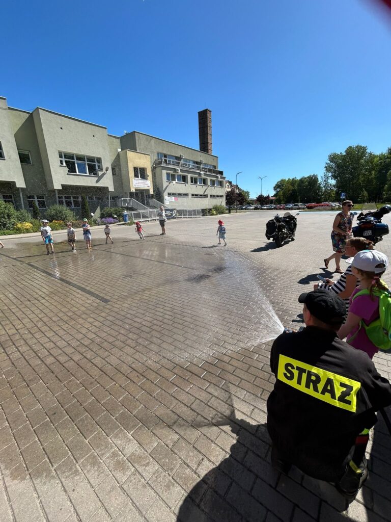 Plac przed budynkiem użyteczności publicznej, na którym strażak w czarnym mundurze z napisem "STRAŻ" polewa wodą dzieci bawiące się na kostce brukowej. Dzieci stoją w rzędzie i wydają się być zadowolone, niektóre z nich mają na sobie czapki chroniące przed słońcem. W tle widać nowoczesny budynek z dużymi oknami i rampą, a także zaparkowane motocykle oraz kilka osób dorosłych. Pogoda jest słoneczna, niebo bezchmurne. Cała scena wygląda na wydarzenie plenerowe, być może pokaz lub festyn strażacki dla dzieci.
