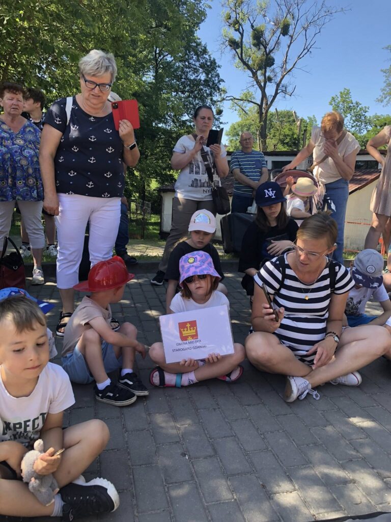 Grupa dzieci i dorosłych na świeżym powietrzu, siedzących i stojących na chodniku. W centrum zdjęcia, na pierwszym planie, siedzi dziewczynka w fioletowym kapeluszu i okularach przeciwsłonecznych, trzymająca kartkę z napisem „GMINA MIEJSKA STAROGARD GDAŃSKI” oraz herbem miasta. Obok niej siedzi kobieta w okularach i w pasiastym t-shircie, trzymająca telefon. Wokół nich siedzi kilkoro dzieci, niektóre mają czapki z daszkiem lub kapelusze, jedno dziecko trzyma pluszaka. W tle stoi kilka dorosłych osób, część z nich robi zdjęcia telefonami. Pogoda jest słoneczna, a wokół rosną drzewa i widać fragmenty budynków. Wszyscy wyglądają na skupionych lub zainteresowanych tym, co się dzieje przed nimi.