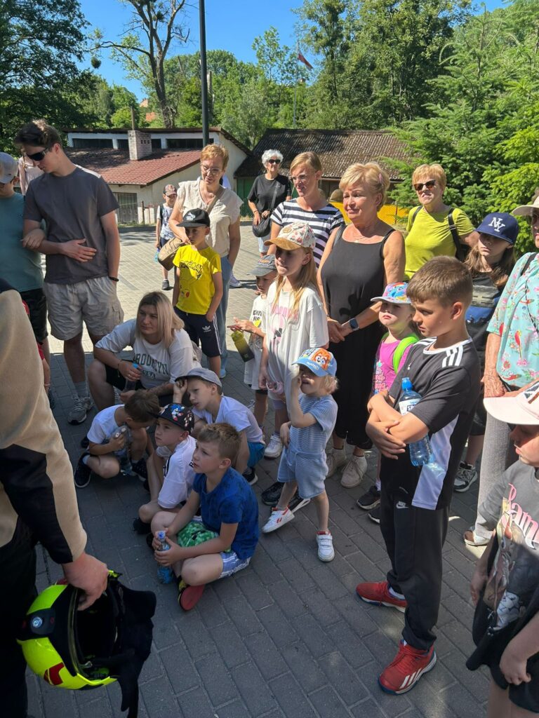 Grupa dzieci i dorosłych na świeżym powietrzu, stojących i siedzących na chodniku. Dzieci, w różnym wieku, są ubrane w letnie ubrania i czapki z daszkiem, niektóre trzymają butelki z wodą. Wśród dorosłych przeważają kobiety, część z nich ma okulary przeciwsłoneczne. W lewym dolnym rogu widoczna jest ręka osoby trzymającej żółty kask strażacki, co sugeruje, że grupa uczestniczy w jakimś pokazie lub spotkaniu edukacyjnym, prawdopodobnie z udziałem strażaka. W tle widać drzewa, budynki i ogrodzenie, a pogoda jest słoneczna. Wszyscy patrzą w jednym kierunku, skupieni na osobie poza kadrem.