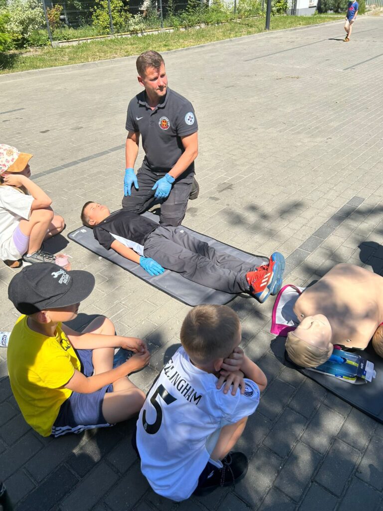 Pokaz pierwszej pomocy na świeżym powietrzu. Na zdjęciu widać mężczyznę w czarnej koszulce z emblematem ratowniczym, który klęczy na macie obok leżącego chłopca. Mężczyzna ma na sobie niebieskie rękawiczki ochronne. Obok nich, na drugiej macie, leży fantom do ćwiczeń resuscytacji krążeniowo-oddechowej. Wokół siedzi kilkoro dzieci, które uważnie obserwują pokaz. Dzieci są ubrane letnio, jedno z nich ma na sobie koszulkę z napisem "BELLINGHAM 5". Całość odbywa się na chodniku, w słoneczny dzień, w pobliżu zieleni i ogrodzenia.