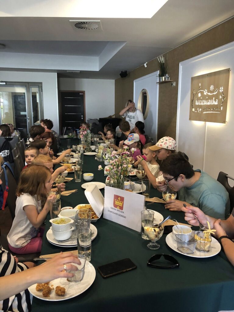 Grupa dzieci i dorosłych siedzących przy długim stole w restauracji. Na stole leży zielony obrus, a na nim stoją talerze z jedzeniem, miseczki z frytkami, szklanki z wodą oraz pucharki z deserem lodowym. Pośrodku stołu stoi tabliczka z herbem i napisem „Gmina Miejska Starogard Gdański”. W tle widać ozdobne kwiaty w wazonach i jasne ściany. Na jednej ze ścian wisi podświetlony szyld z napisem „Boccianka”. Dzieci jedzą, rozmawiają i wyglądają na zadowolone. Pomieszczenie jest jasne i nowoczesne.
