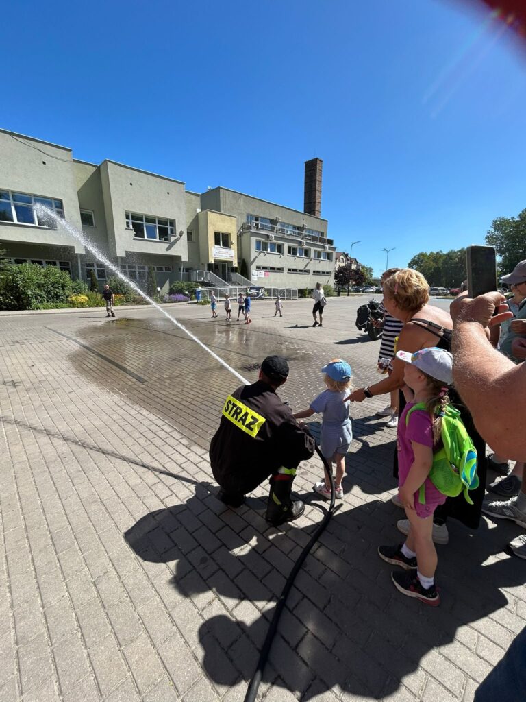 Plac przed budynkiem użyteczności publicznej, prawdopodobnie szkołą lub domem kultury, w słoneczny dzień. Na pierwszym planie strażak w czarnym mundurze z żółtym napisem "STRAŻ" klęczy obok małego dziecka w niebieskiej czapeczce i jasnym ubranku. Razem trzymają wąż strażacki, z którego leci silny strumień wody w kierunku placu. Wokół nich stoi grupa dzieci i dorosłych, niektórzy robią zdjęcia telefonami. W tle widać więcej dzieci bawiących się na placu, a także zieleń i fragmenty ulicy. Całość sprawia wrażenie pokazu lub festynu strażackiego dla dzieci.
