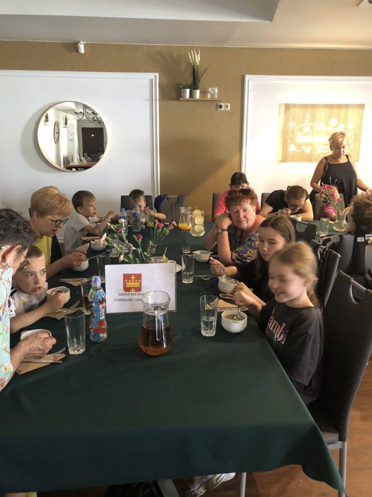 Grupa osób siedzących przy długim stole w restauracji lub stołówce. Przy stole siedzą głównie dzieci oraz kilka dorosłych kobiet. Wszyscy jedzą z białych miseczek, na stole stoją dzbanki z napojami (herbata i napój cytrynowy), szklanki z wodą oraz butelka wody mineralnej. Na środku stołu stoi tabliczka z napisem „GMINA MIEJSKA STARGARD GDAŃSKI” oraz żółtą koroną na czerwonym tle. Stół przykryty jest ciemnozielonym obrusem, a na środku stoją wazony z kwiatami. W tle widać lustro, roślinę na półce i jasne ściany. W pomieszczeniu panuje spokojna, rodzinna atmosfera.