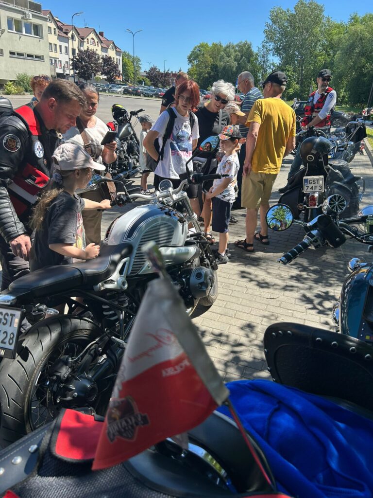 Grupa ludzi zgromadzona wokół motocykli na chodniku w słoneczny dzień. W centrum zdjęcia znajduje się kilka dzieci i dorosłych, którzy oglądają i dotykają srebrny motocykl. Wśród dorosłych są osoby w kamizelkach motocyklowych, a także kobieta z siwymi włosami w okularach przeciwsłonecznych. Na pierwszym planie widać fragment motocykla z małą polską flagą i logotypem Harley-Davidson. W tle stoją kolejne motocykle oraz osoby, a po lewej stronie widoczne są budynki mieszkalne i zaparkowane samochody. Pogoda jest bardzo słoneczna, niebo bezchmurne, a drzewa w tle są zielone.