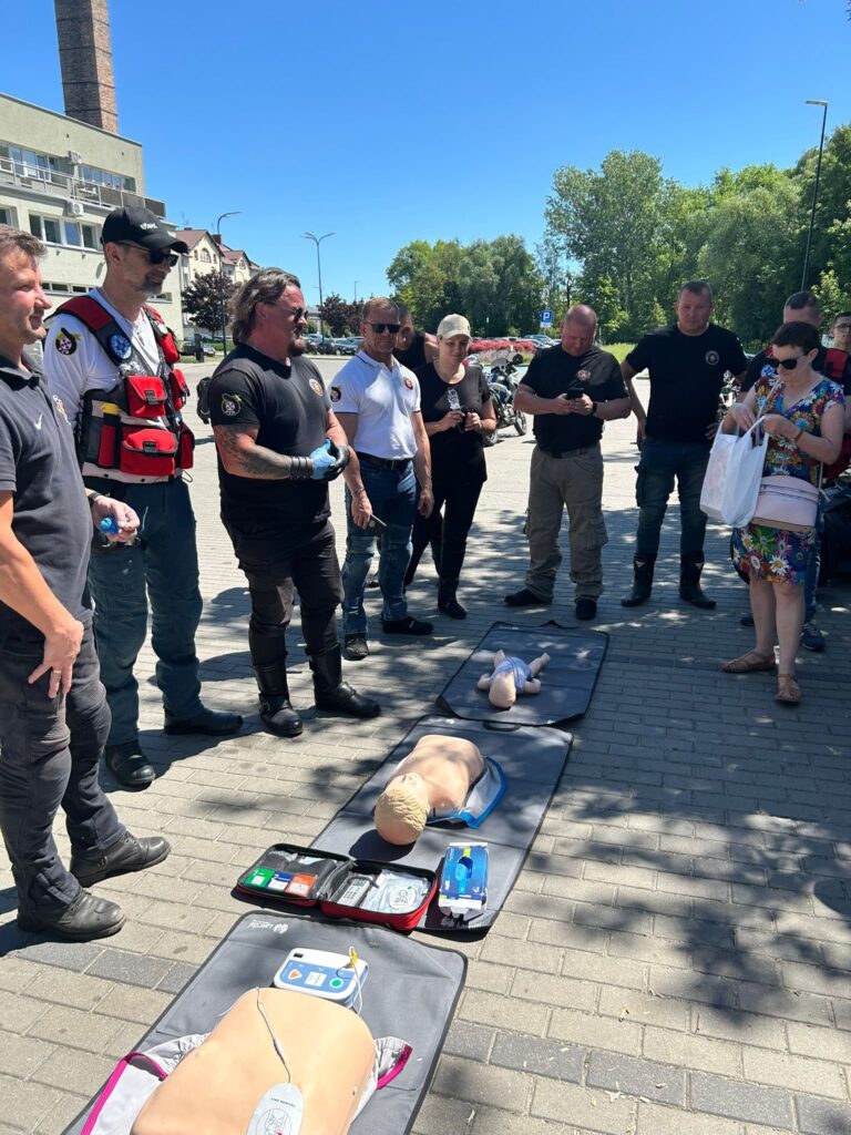 Grupa ludzi stojąca na zewnątrz w półkolu wokół trzech fantomów do nauki resuscytacji krążeniowo-oddechowej (RKO), ułożonych na matach na chodniku. Na fantomach widoczne są podłączone elektrody treningowych defibrylatorów AED oraz otwarte zestawy ratownicze. Uczestnicy mają na sobie różnorodne ubrania – część osób nosi czarne koszulki z emblematami, jedna osoba ma na sobie biały t-shirt, a inna czerwoną kamizelkę ratowniczą. W tle widoczne są budynki, drzewa oraz zaparkowane motocykle. Pogoda jest słoneczna, niebo bezchmurne. Wygląda na to, że odbywa się szkolenie z pierwszej pomocy.