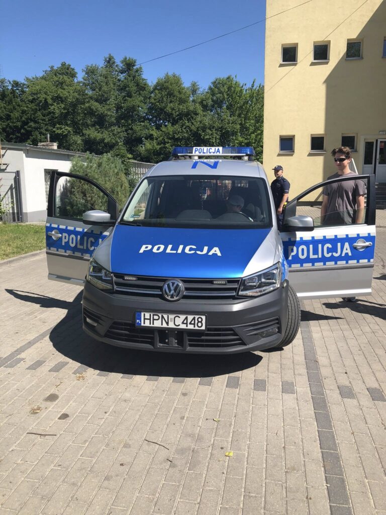 Radiowóz policyjny marki Volkswagen stojący na podjeździe przed budynkiem. Samochód ma otwarte obie przednie drzwi, na masce i drzwiach widnieje napis „POLICJA”. Na dachu znajduje się niebieski sygnał świetlny z napisem „POLICJA”. Przed samochodem stoi młody mężczyzna w okularach przeciwsłonecznych, a obok budynku stoi policjant w mundurze. W tle widoczne są drzewa, ogrodzenie i fragment białego budynku. Pogoda jest słoneczna, niebo bezchmurne.