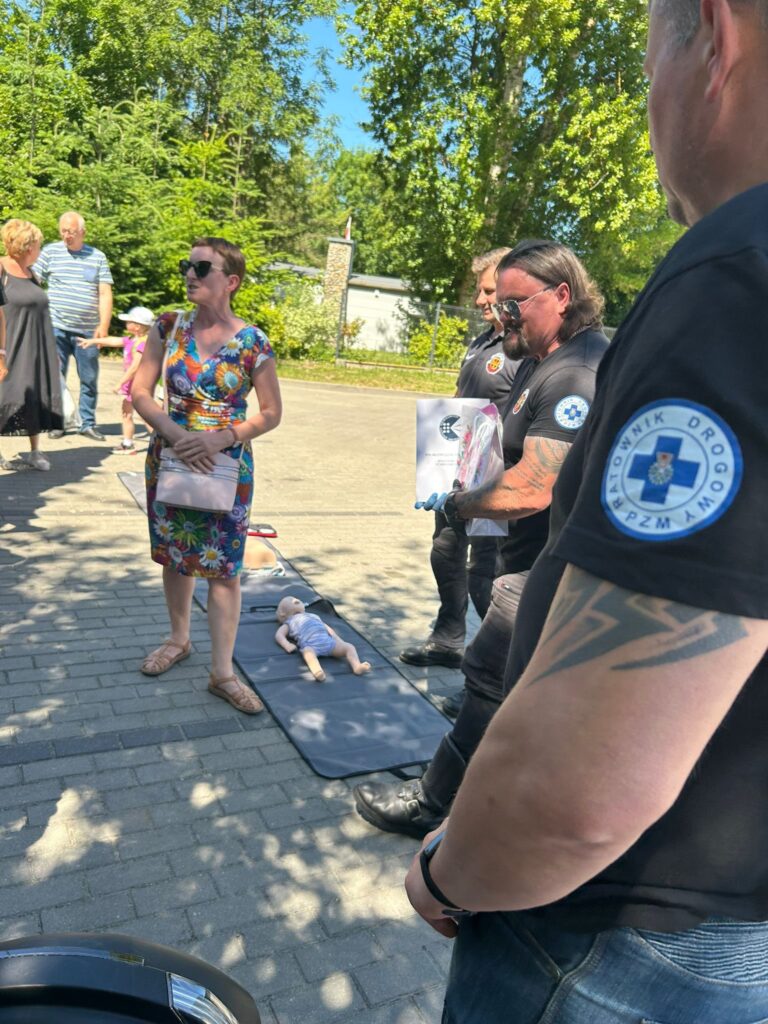Grupa ludzi na świeżym powietrzu, stojących na chodniku w cieniu drzew. Na pierwszym planie widoczny jest mężczyzna z tatuażem na ramieniu i naszywką „Ratownik Drogowy” na rękawie. Obok niego stoi kobieta w kolorowej sukience w kwiaty, z jasną torebką przewieszoną przez ramię. Na ziemi, na rozłożonej macie, leżą dwa manekiny do nauki pierwszej pomocy – jeden w rozmiarze niemowlęcia, drugi większy. W tle stoją inni ludzie, w tym mężczyzna w okularach przeciwsłonecznych i z długimi włosami, trzymający torbę lub pakiet. Scena wygląda na pokaz lub szkolenie z udzielania pierwszej pomocy, prowadzone na zewnątrz w słoneczny dzień.