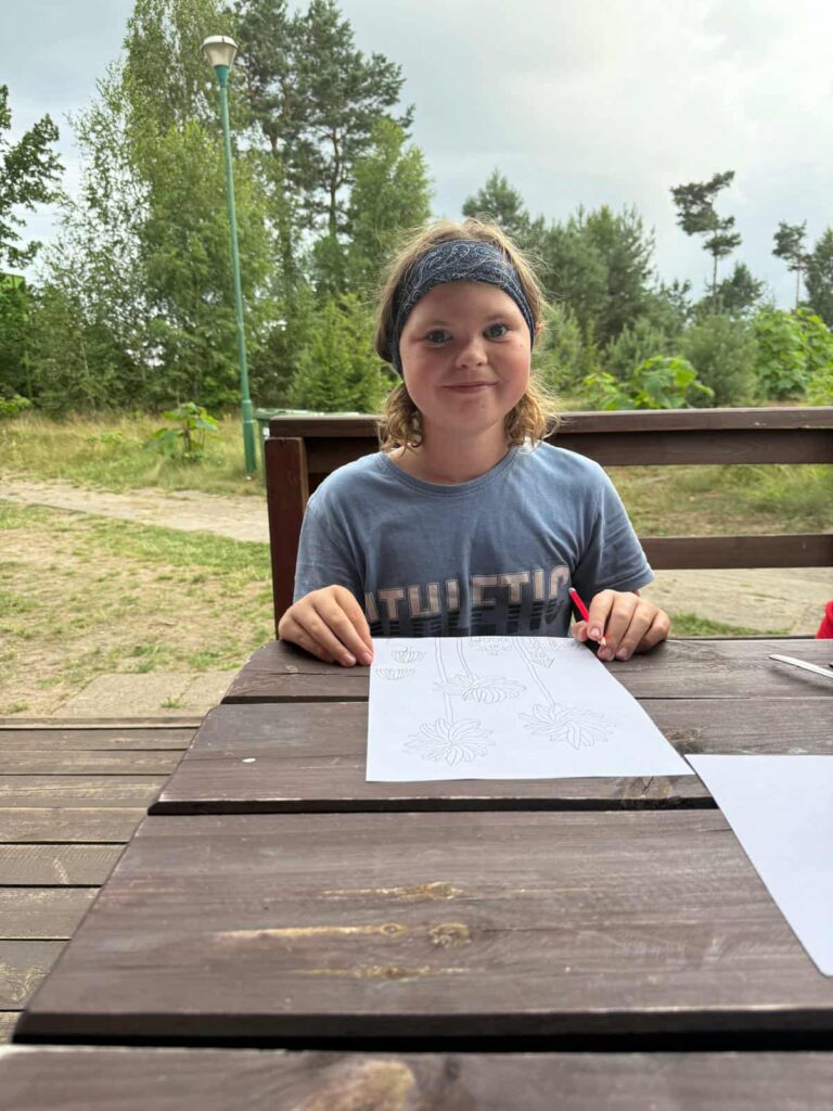 Na zdjęciu widzimy dziecko siedzące przy drewnianym stole na świeżym powietrzu, skupione na twórczym zajęciu. Ubrane w niebieską opaskę i koszulkę z napisem „THLETIC”, trzyma w dłoni czerwoną kredkę, kolorując kartkę z konturami kwiatów i liści. Otoczenie jest naturalne — w tle widać drzewa, krzewy i ścieżkę z ziemi, co sugeruje, że scena rozgrywa się w parku lub ogrodzie. Atmosfera jest spokojna i sprzyja kreatywności, a dziecko wydaje się całkowicie pochłonięte artystyczną aktywnością.