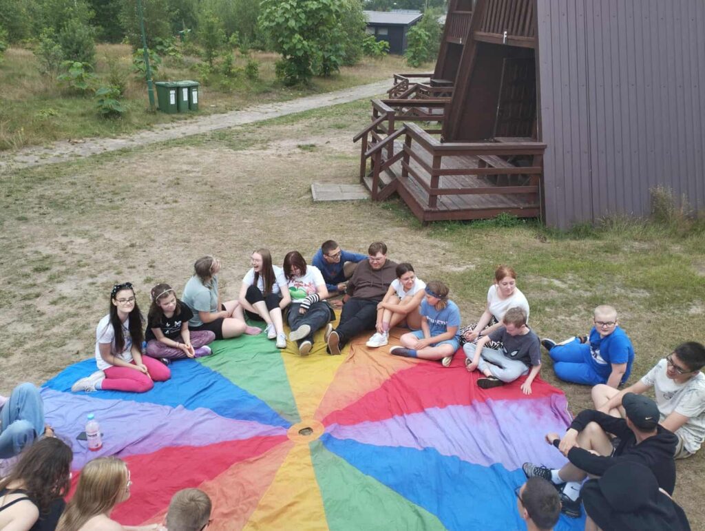 Grupa osób siedzi w kręgu na kolorowej macie przypominającej spadochron, rozłożonej na trawie w otoczeniu natury. Każdy segment maty ma inny kolor — czerwony, niebieski, zielony, żółty, fioletowy — zbiegające się w centralnym punkcie, co tworzy efekt tęczowego wiru. Uczestnicy, ubrani swobodnie, wyglądają na zaangażowanych w wspólną aktywność — może to być warsztat, gra integracyjna albo po prostu chwila rozmowy. W tle widać drewniane domki, ścieżkę z ziemi i bujną roślinność, co sugeruje, że scena rozgrywa się na obozie lub podczas plenerowego spotkania. Całość emanuje atmosferą wspólnoty, luzu i letniego wypoczynku.