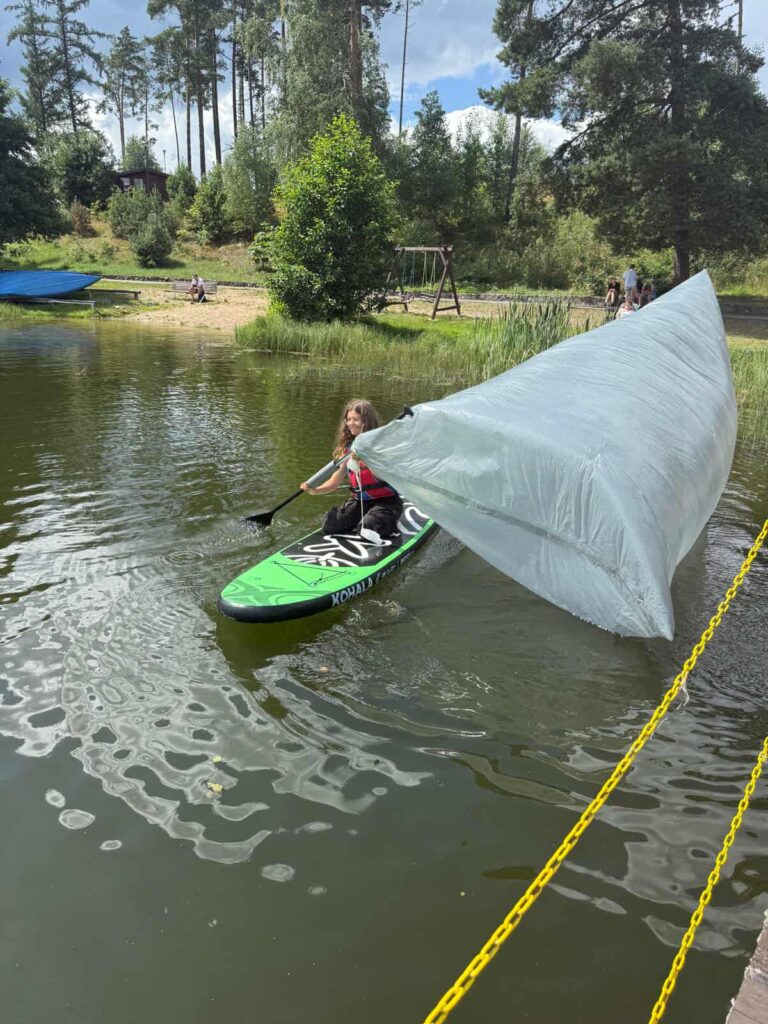 Na spokojnej tafli jeziora, w otoczeniu zieleni i piasku, jedna osoba balansuje na zielonej desce typu paddleboard z napisem „KONA”. W rękach trzyma wiosło, do którego przymocowana jest duża, przezroczysta płachta — niczym prowizoryczny żagiel — łapiąca wiatr i nadająca całej scenie niecodzienny, eksperymentalny charakter. Płachta unosi się w powietrzu, sugerując, że natura właśnie współpracuje z pomysłowością człowieka. W tle widać huśtawkę, niebieską łódkę na brzegu i żółte łańcuchy wyznaczające strefę bezpieczeństwa. To obraz kreatywności, zabawy i kontaktu z żywiołami — gdzie sport wodny spotyka się z improwizacją.