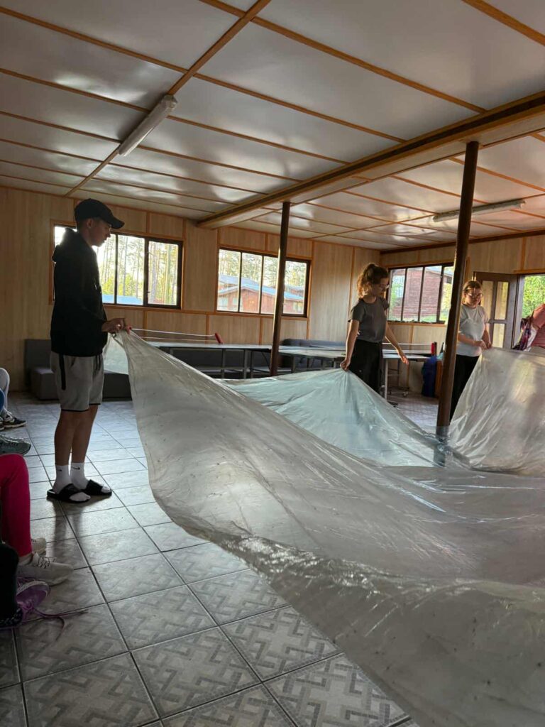 W przestronnym pomieszczeniu o geometrycznej podłodze i drewnianych ścianach grupa osób wspólnie rozkłada dużą, przezroczystą folię. Naturalne światło wpada przez szerokie okna, a w tle widać kanapy, miotłę i inne elementy codziennego wyposażenia, co nadaje miejscu domowy, wspólnotowy charakter. Cała scena emanuje atmosferą współpracy — jakby uczestnicy przygotowywali salę na wydarzenie, dekorację lub kreatywną aktywność. To moment skupienia, zaangażowania i wspólnego działania, gdzie każdy gest ma znaczenie, a przestrzeń zmienia się dzięki ludzkiej energii.