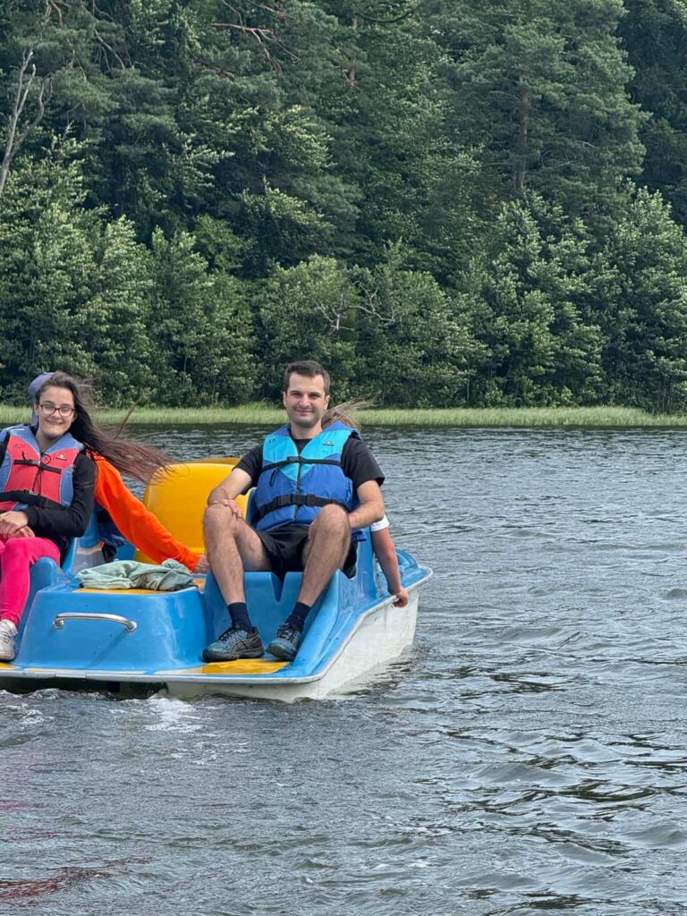 Na spokojnym jeziorze, otoczonym gęstym lasem, dwie osoby siedzą na niebiesko-białej łódce wodnej, delektując się chwilą relaksu. Ubrane w kamizelki ratunkowe — jedna czerwona, druga niebieska — wyglądają na zanurzone w rozmowie lub po prostu w ciszy natury. Osoba po lewej ma długie włosy, jaskrawe różowe spodnie i czerwoną kurtkę, a po prawej — krótkie włosy, czarną koszulkę i niebieską kamizelkę. Woda delikatnie faluje, a tło tworzy soczysta zieleń drzew i roślinności. To moment wyciszenia, bezpieczeństwa i bliskości z naturą — idealny przykład letniego odpoczynku w dobrym towarzystwie.