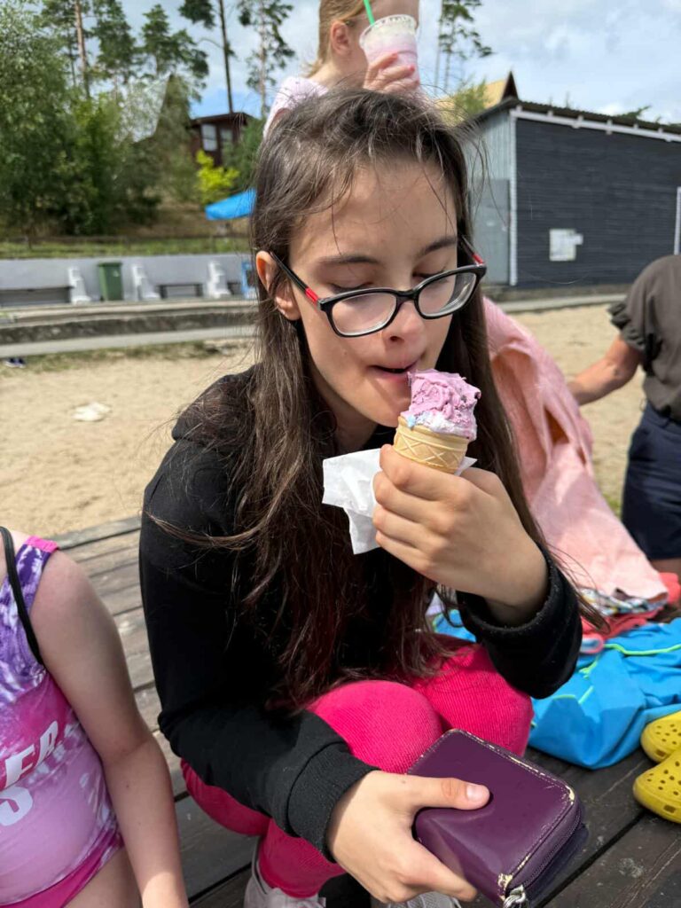 Na drewnianym podwyższeniu, tuż przy piaszczystym terenie, młoda osoba delektuje się różowym lodem w wafelku, trzymając w drugiej ręce fioletowy portfel. Ubrana w czarną bluzę i jaskrawe różowe spodnie, z okularami na nosie, wygląda na zrelaksowaną i zadowoloną. W tle widać innych uczestników — dziecko z zielonym napojem i osobę w fioletowej bluzie — a także budynek o ciemnej elewacji, drzewa oraz rozłożone ręczniki. Cała scena tchnie letnim luzem, chwilą odpoczynku i prostą radością z małych przyjemności.