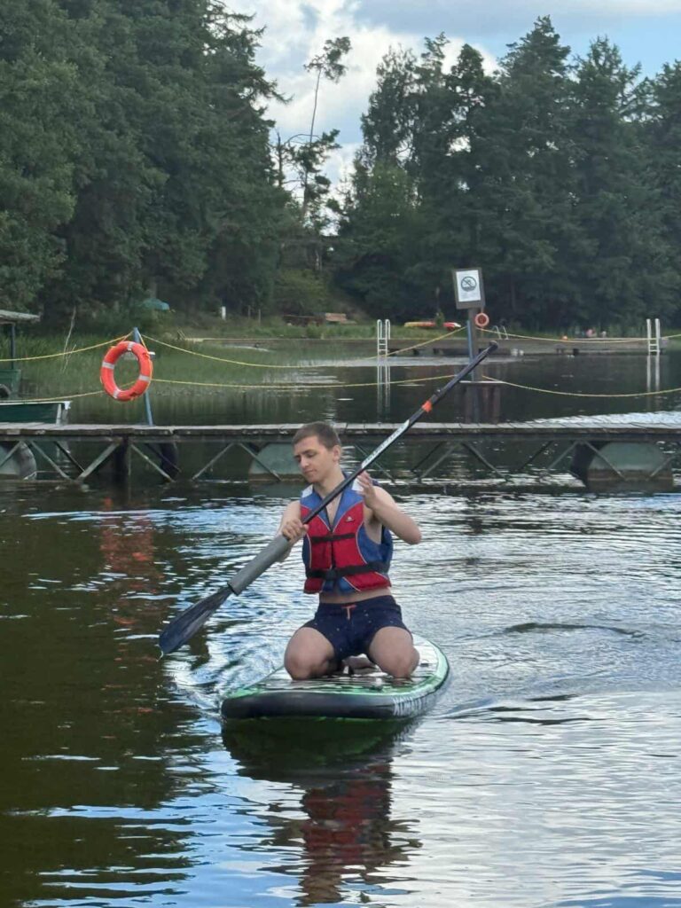 Osoba w czerwono‑niebieskiej kamizelce klęczy na zielonej desce „ATHLETIC” i wiosłuje po spokojnym jeziorze. W tle pomost z kołem ratunkowym, drzewa i częściowo zachmurzone niebo.