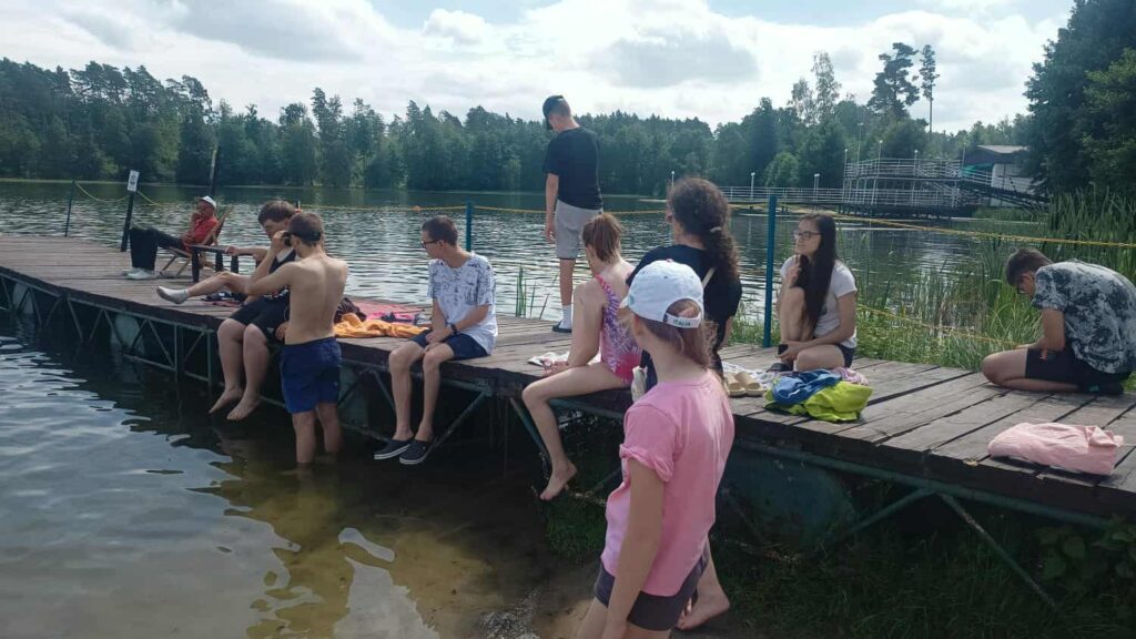 Grupa dzieci i nastolatków spędza czas na drewnianym pomoście nad jeziorem, otoczonym drzewami, przy częściowo zachmurzonym niebie.