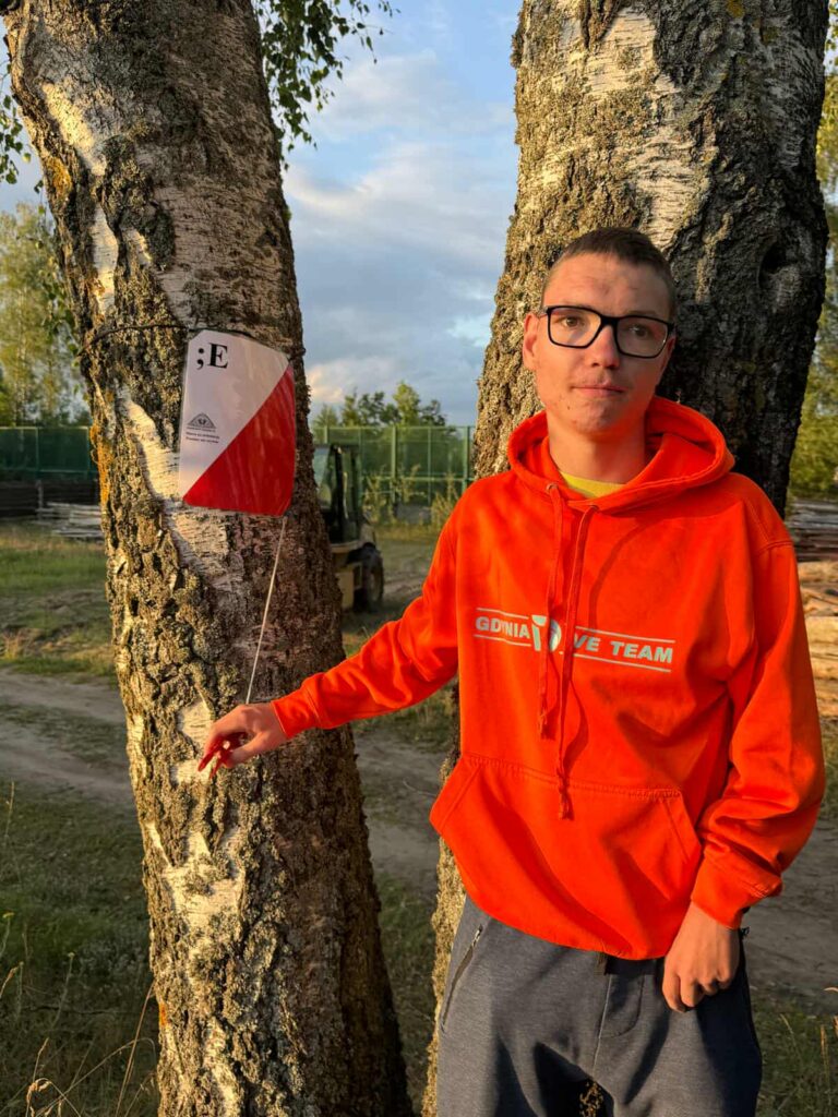 Osoba ubrana w pomarańczową bluzę z napisem „ODINA LOVE TEAM” stoi obok drzewa, wskazując na małą trójkątną flagę przymocowaną do pnia. Flaga ma czerwono-białe barwy i widnieje na niej litera „E”. W tle widać nieutwardzoną ścieżkę, zielone ogrodzenie, wózek widłowy oraz drzewa, co sugeruje, że zdjęcie zostało wykonane w plenerze — prawdopodobnie w lesie lub parku. Elementy takie jak flaga mogą wskazywać na aktywność terenową, np. bieg na orientację lub grę zespołową.