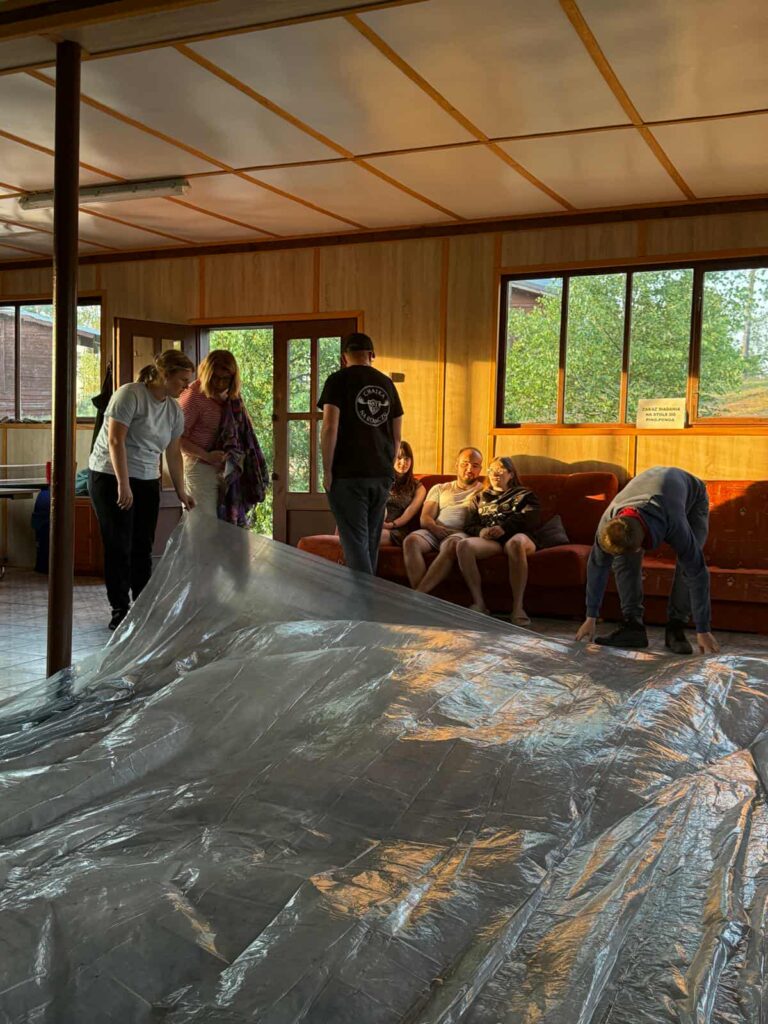 W jasnym, drewnianym pomieszczeniu z dużymi oknami grupa osób wspólnie rozkłada przezroczystą folię lub plandekę na podłodze. Naturalne światło wpada przez okna, nadając wnętrzu ciepły klimat. W tle kilka osób odpoczywa na czerwonej kanapie, a jedna z nich ma na sobie czarną koszulkę z okrągłym logo na plecach. Na oknie widnieje kartka z napisem „PLEASE DO NOT SIT ON THE BACK OF THE COUCH”. Całość sprawia wrażenie przygotowań do jakiegoś wydarzenia lub aktywności zespołowej, w przyjaznej i nieformalnej atmosferze.