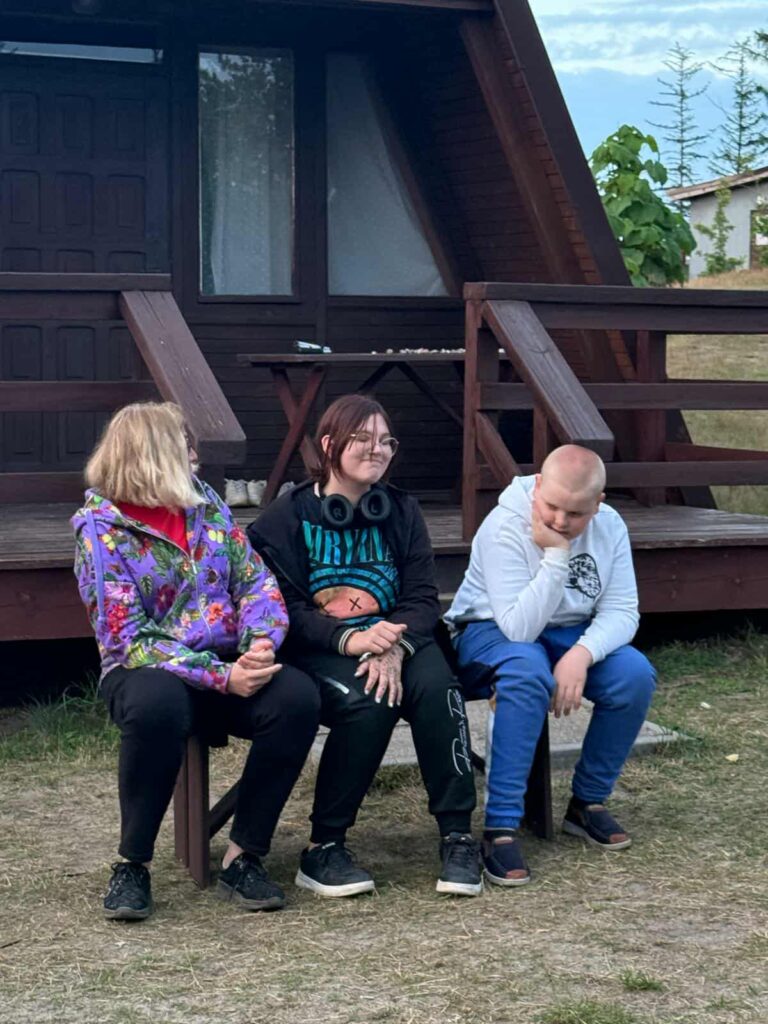 Trójka osób siedzi na ławce przed drewnianą chatką z trójkątnym dachem, która wygląda jak przytulne schronienie wśród natury. Chatka ma mały ganek ze schodkami i stolik z krzesłami, sugerując miejsce do odpoczynku lub wspólnych rozmów. Osoba po lewej ma na sobie kolorową kurtkę w żywe wzory, środkowa nosi czarną bluzę z logo zespołu Nirvana i słuchawki na szyi, a trzecia osoba ubrana jest w jasnoszarą bluzę i niebieskie spodnie. Tło tworzy zielona trawa i drzewa, co wskazuje na spokojne, wiejskie otoczenie — być może obóz, weekendowy wypad lub miejsce integracji.