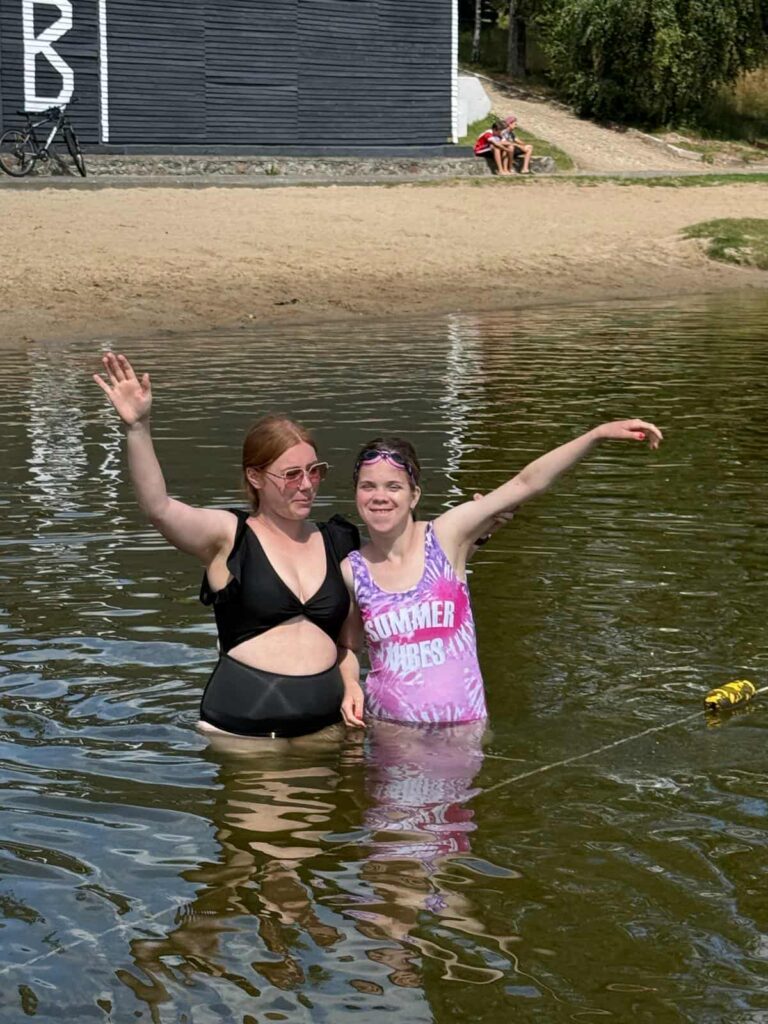 Dwie osoby stoją w płytkiej wodzie przy brzegu jeziora lub morza, ciesząc się letnią aurą. Jedna z nich, ubrana w czarny strój kąpielowy, macha ręką w stronę aparatu, podczas gdy druga, w różowo-fioletowym kostiumie z napisem „SUMMER VIBES”, rozkłada ramiona w geście radości. W tle widać piaszczystą plażę, budynek z literą „B”, rower oraz kilka osób odpoczywających na piasku. Cała scena emanuje beztroską atmosferą wakacyjnego relaksu i wspólnej zabawy.