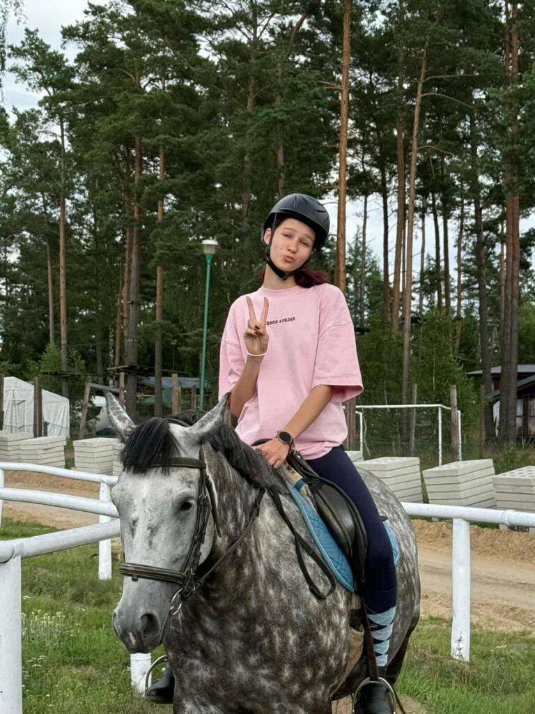 Osoba siedzi na siwym koniu o cętkowanej sierści, ubrana w jasnoróżowy, luźny T-shirt, ciemne bryczesy i wysokie buty do jazdy konnej. Na głowie ma czarny kask, a dłonią pokazuje znak pokoju, sprawiając wrażenie zrelaksowanej i zadowolonej. W tle widać ogrodzony teren do jazdy konnej z białymi barierkami, ścieżkę z ziemi oraz wysokie sosny, co wskazuje na otwarte, naturalne otoczenie — prawdopodobnie szkółkę jeździecką lub rekreacyjny ośrodek. W oddali dostrzec można bramkę do piłki nożnej i niewielki budynek, co dodaje miejscu wielofunkcyjnego charakteru.