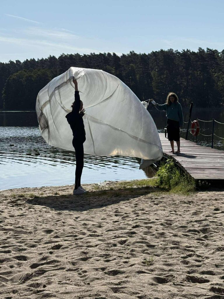 Dwójka osób znajduje się nad jeziorem — jedna stoi na piaszczystym brzegu, druga na drewnianym pomoście. Trzymają wspólnie dużą, półprzezroczystą tkaninę lub siatkę, która unosi się w powietrzu, tworząc efektowną, niemal eteryczną formę. Jezioro jest spokojne, otoczone gęstym lasem, a niebo czyste, co nadaje całej scenie atmosferę harmonii i lekkości. Ruch materiału, światło i naturalne otoczenie tworzą wizualnie intrygujący moment — jakby uchwycono fragment tańca z wiatrem.