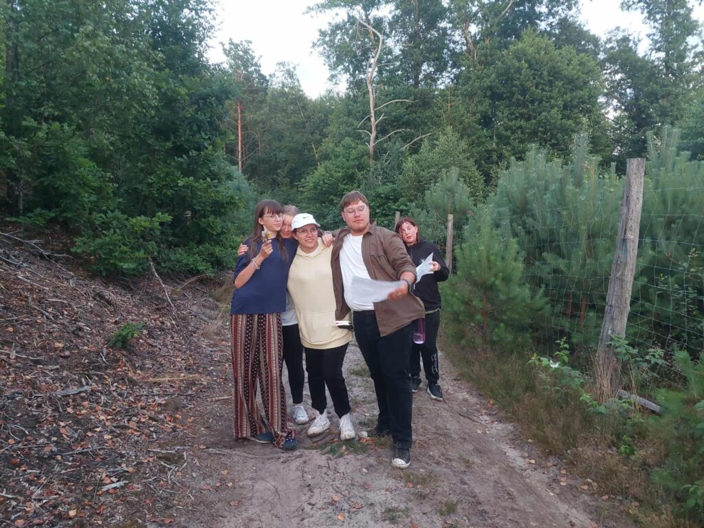 Cztery osoby stoją na leśnej ścieżce otoczonej gęstą zielenią — wysokimi drzewami i krzewami. Jedna z nich trzyma mapę lub kartkę, co sugeruje, że grupa może być w trakcie wędrówki lub gry terenowej. Ubrani są swobodnie, odpowiednio do aktywności na świeżym powietrzu. Po prawej stronie ścieżki ciągnie się ogrodzenie z drewnianych słupków i drutu. Cała scena rozgrywa się w świetle dnia, w spokojnym, naturalnym otoczeniu, które sprzyja eksploracji i wspólnemu spędzaniu czasu.