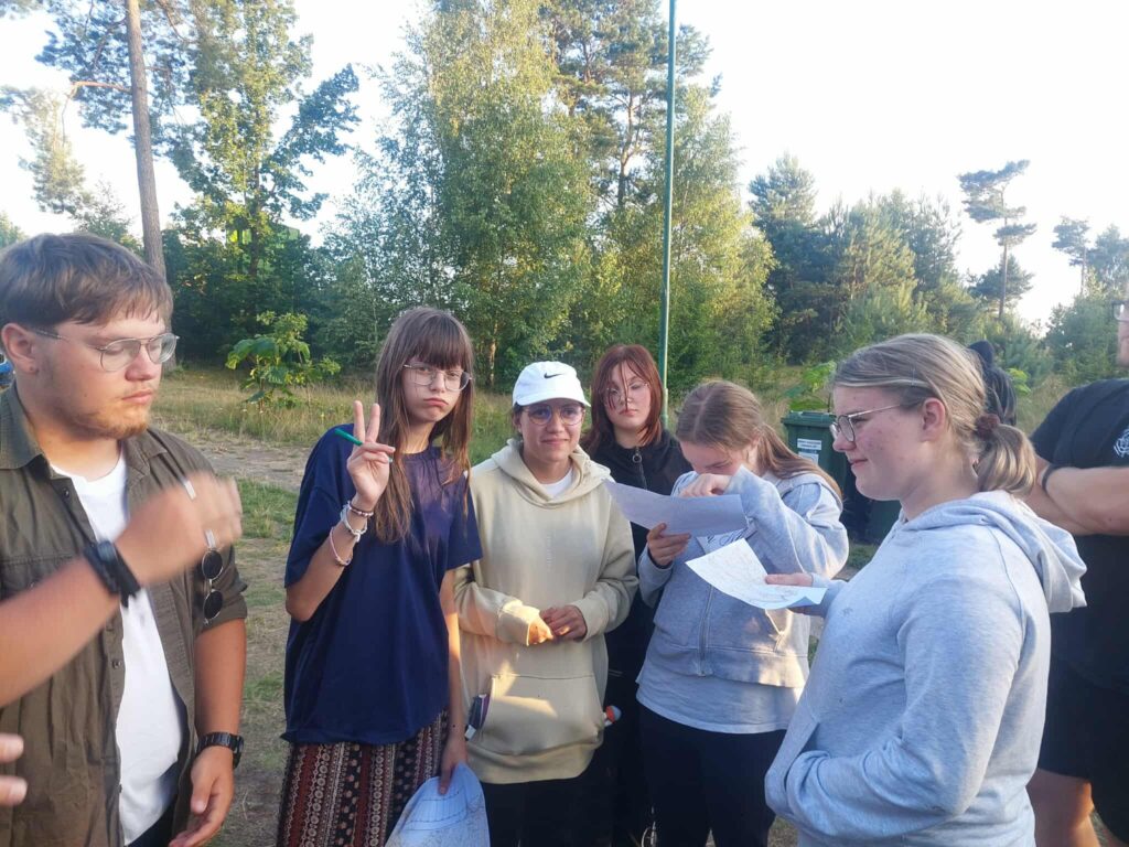 Siedmioro młodych osób stoi razem w otoczeniu zieleni, w miejscu przypominającym leśną polanę lub park. W rękach trzymają kartki, co sugeruje, że uczestniczą w zadaniu grupowym — może to być gra terenowa, quiz albo wspólne rozwiązywanie zagadek. Jedna osoba pokazuje znak pokoju, inne są skupione na kartkach lub rozmawiają. Ubrani są swobodnie, a światło — miękkie i ciepłe — wskazuje na poranek lub późne popołudnie. Cała scena oddaje klimat współpracy, luzu i dobrej energii w naturalnym otoczeniu.