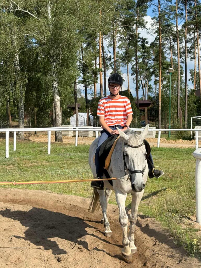 Osoba w czerwono-białej koszulce w paski, ciemnych spodniach i czarnym kasku jeździeckim dosiada białego konia na piaszczystym terenie do jazdy konnej. Koń wyposażony jest w siodło i uzdę, a cała scena rozgrywa się w ogrodzonym obszarze otoczonym wysokimi drzewami. W tle widać budynki oraz bramkę do piłki nożnej, co sugeruje, że miejsce pełni funkcję rekreacyjną. Jasne światło dnia i spokojna atmosfera nadają zdjęciu lekkości i poczucia harmonii między człowiekiem a zwierzęciem.