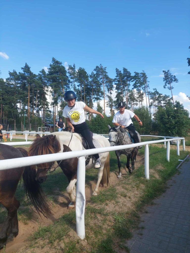 Dwójka osób jedzie konno wzdłuż ogrodzonej ścieżki na świeżym powietrzu. Jeden z jeźdźców dosiada białobrązowego konia i ma na sobie biały T-shirt z żółtą buźką, drugi jedzie na siwym koniu w zwykłej białej koszulce. Oboje mają kaski i wyglądają na zrelaksowanych, jakby uczestniczyli w zabawie lub ćwiczeniu wymagającym współpracy — ich gesty sugerują, że próbują sięgnąć do siebie lub zsynchronizować ruchy. Otoczenie to zielona przestrzeń z trawnikiem, chodnikiem i wysokimi sosnami pod błękitnym niebem, co nadaje scenie pogodny, letni klimat.