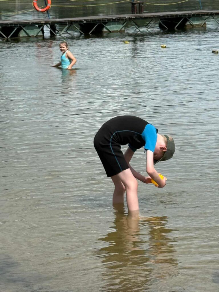Dwójka dzieci bawi się w płytkiej wodzie przy brzegu jeziora lub plaży. Jedno z nich, ubrane w czarno-niebieski kombinezon i czapkę, pochyla się nad wodą, badając coś z pomocą żółtego przedmiotu — może to zabawka, pojemnik lub narzędzie do eksploracji. Drugie dziecko, w jasnoniebieskim stroju kąpielowym, stoi dalej w wodzie i patrzy w stronę aparatu. W tle widać pomost z kołem ratunkowym i pływającymi znacznikami, a spokojna tafla wody oraz czyste niebo tworzą sielankową scenerię. Całość oddaje klimat dziecięcej ciekawości, zabawy i kontaktu z naturą.
