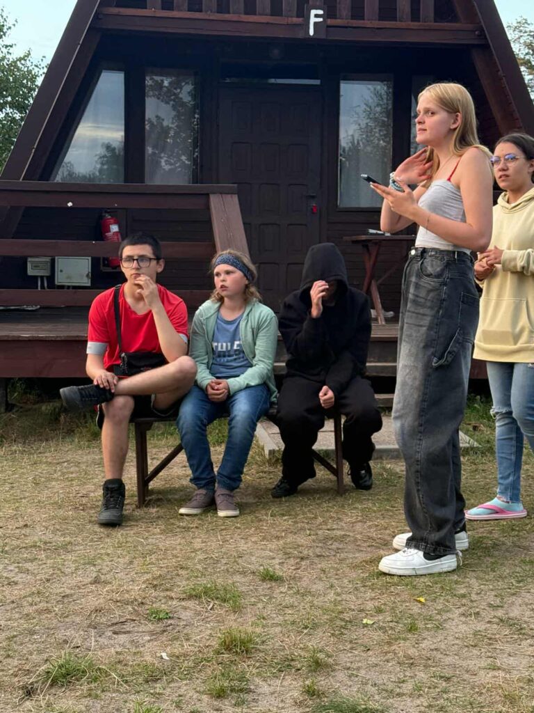 Pięć osób znajduje się przed drewnianą chatką z trójkątnym dachem, nad której drzwiami widnieje litera „F”. Na werandzie wisi gaśnica, a obok stoi ławka. Grupa rozsiadła się lub stoi na trawie — jedna osoba prezentuje coś, trzymając telefon lub małe urządzenie, inna, w czerwonej koszulce i okularach, trzyma aparat fotograficzny. Pozostali słuchają, rozmawiają lub po prostu odpoczywają, a jedna z osób ma na głowie kaptur, który zasłania twarz. Całość tworzy obraz swobodnego spotkania w plenerze, być może w ramach obozu, warsztatów lub wspólnego projektu.