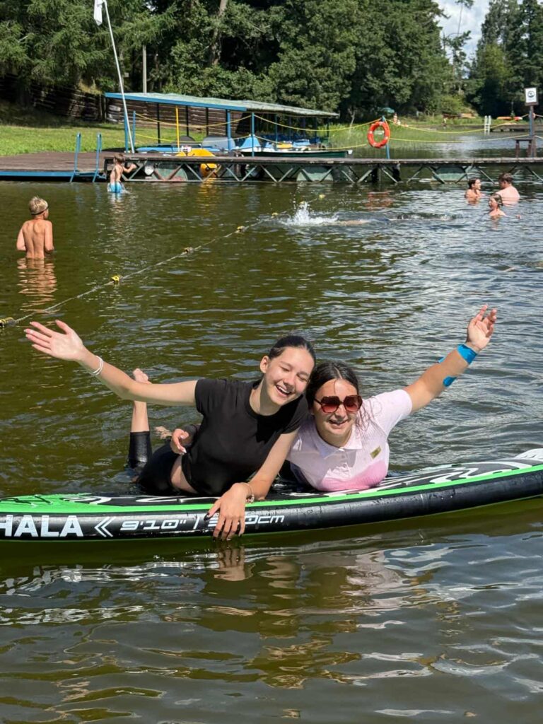 Dwójka osób siedzi na desce do paddleboardingu na spokojnym jeziorze, uśmiechając się i machając do aparatu. Jedna z nich ma na sobie czarną koszulkę, druga jasnoróżową i okulary przeciwsłoneczne. W tle widać pomost z kołem ratunkowym, a wokół nich inni uczestnicy kąpią się i bawią w wodzie. Dalej w tle dostrzec można drzewa i budynek, co wskazuje na obozowy lub rekreacyjny charakter miejsca. Cała scena emanuje radością, luzem i letnią energią — to moment beztroskiej zabawy i wspólnego relaksu na łonie natury.