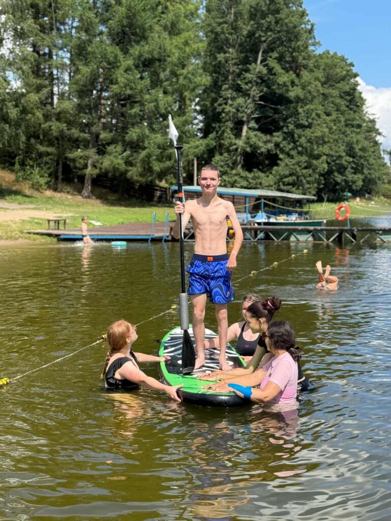 Pięć osób spędza aktywnie czas nad jeziorem. Na środku uwagi znajduje się młody mężczyzna stojący na desce do paddleboardingu, trzymający wiosło pionowo niczym włócznię — ubrany w niebieskie kąpielówki z wzorem. Wokół niego, w wodzie, znajdują się cztery inne osoby — trzy kobiety i jeden mężczyzna — którzy wspierają go lub wchodzą z nim w interakcję. W tle widać pomost, osoby kąpiące się i spacerujące, a całość otoczona jest bujną zielenią drzew. Scena emanuje energią, zabawą i wspólnotą — to moment letniej beztroski i przyjacielskiej więzi.