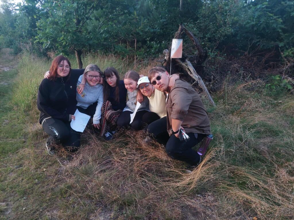 Sześć osób siedzi lub kuca blisko siebie na trawie w otoczeniu drzew i wysokiej roślinności. W tle znajduje się niewielka drewniana konstrukcja z przymocowanym biało-pomarańczowym znacznikiem, przypominającym punkt kontrolny z biegu na orientację. Jedna osoba trzyma kartkę — prawdopodobnie mapę lub instrukcję — co sugeruje, że grupa bierze udział w terenowej grze, zadaniu przygodowym lub aktywności na świeżym powietrzu. Cała scena oddaje klimat wspólnego działania, skupienia i przyjemności z eksplorowania natury.