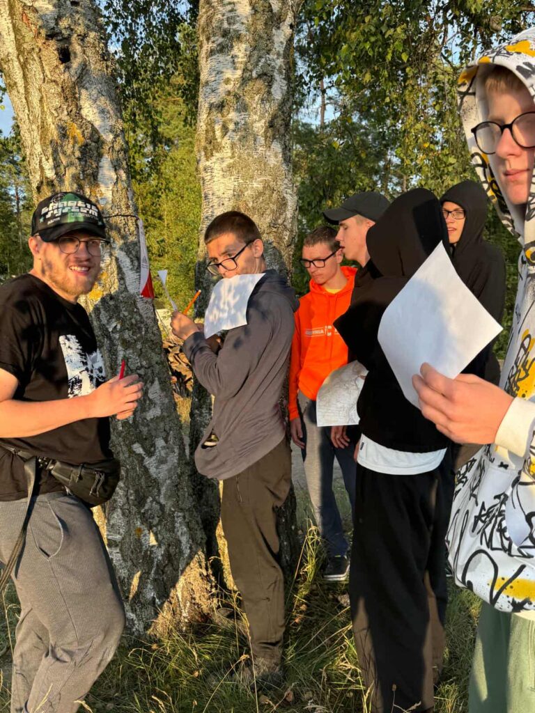 Siedem osób stoi wśród brzóz, skupionych wokół aktywności z papierem i długopisami. Jedna osoba przyciska kartkę do pnia drzewa, jakby robiła odbitkę kory lub notatki terenowe, inna uśmiecha się do aparatu z długopisem w ręku. Ubrani są swobodnie — bluzy, kurtki, okulary — a promienie słońca przebijające się przez liście nadają scenie ciepły, poranny lub popołudniowy klimat. Całość przypomina moment z warsztatów przyrodniczych, gry terenowej albo wspólnego odkrywania świata w rytmie natury.