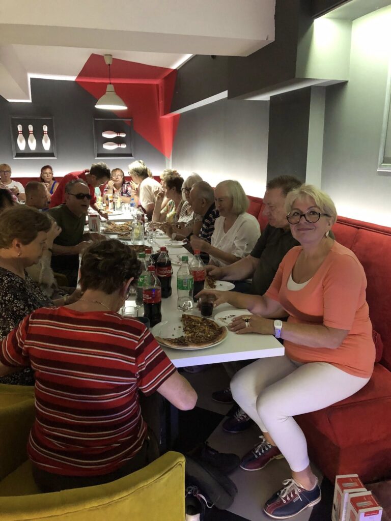 Grupa osób siedzi przy długim stole w restauracji z czerwonymi i szarymi ścianami; na stole leżą pizze, butelki Coca-Coli i Sprite oraz kieliszki z winem; w tle widać dekoracje w formie kręgli i wiszące lampy tworzące przytulną atmosferę.