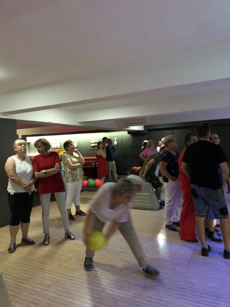 Osoba w ruchu rzucająca żółtą kulę do kręgli, otoczona grupą obserwatorów; w tle widać drewniane tory, kręgle i urządzenia zwrotu kul.