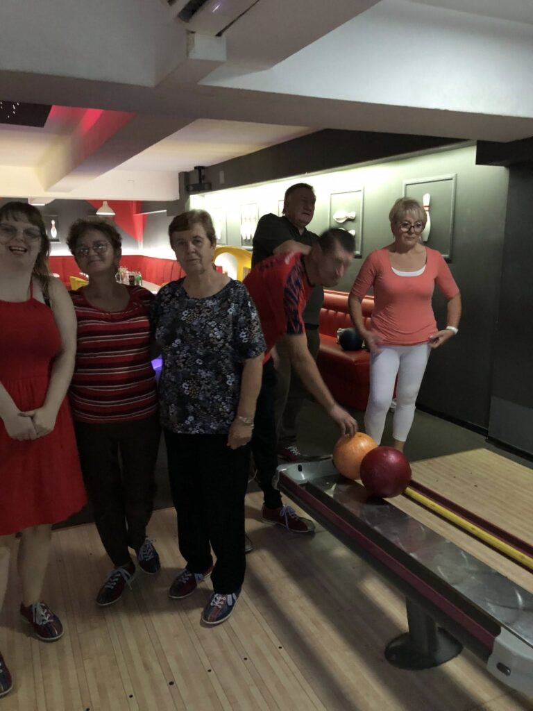 Sześć osób stoi na drewnianej nawierzchni torów do kręgli; jedna osoba pochyla się nad zwrotnikiem kul, wybierając pomarańczową lub czerwoną kulę, a pozostali stoją, pozując do zdjęcia; w tle czerwone siedziska i dekoracje na ścianie z motywami kręglarskimi; wszyscy mają buty do kręgli; oświetlenie jest przyciemnione z czerwonymi i białymi akcentami.